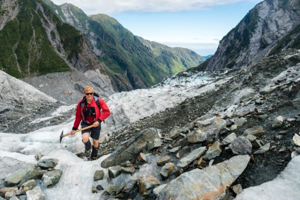 A person is trekking on a glacier amidst rocky terrain, equipped with a backpack and hiking poles, with rugged, green mountains stretching into the distance.