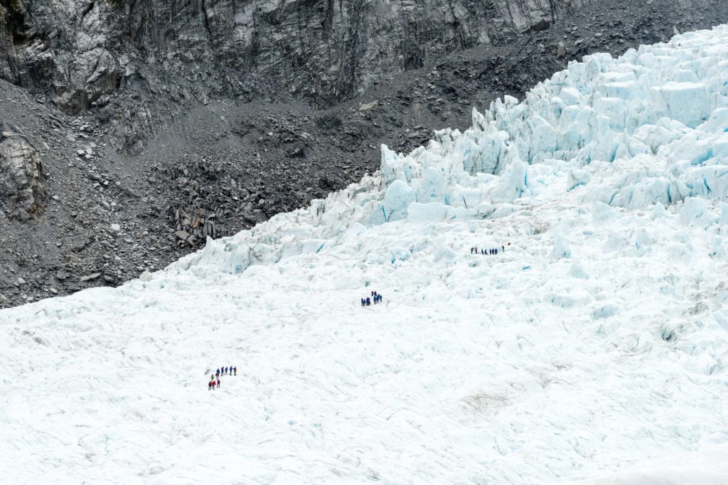 Several groups of people are trekking across a vast glacier, surrounded by rugged terrain. The ice formation's textures and crevasses indicate a dynamic landscape.