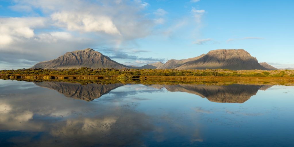 This image features a serene landscape with two prominent mountains reflected in the still waters of a lake under a partly cloudy blue sky.