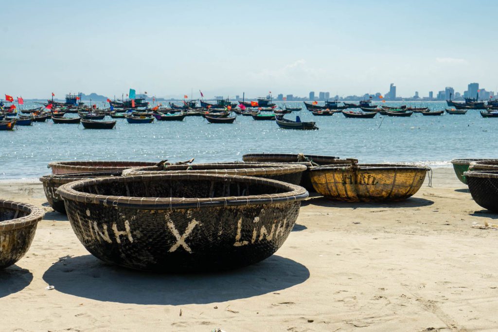 Round basket boats on a sandy beach with numerous traditional fishing boats anchored at sea. City skyline under a clear blue sky in the background.