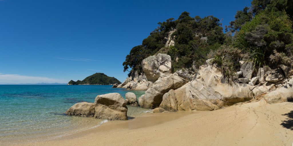 This image features a serene beach with golden sand, clear turquoise waters, and a backdrop of lush greenery and rocky formations under a blue sky.