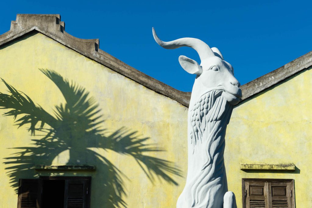 A large goat sculpture against a yellow building with strong palm tree shadows cast on the wall under a clear blue sky.