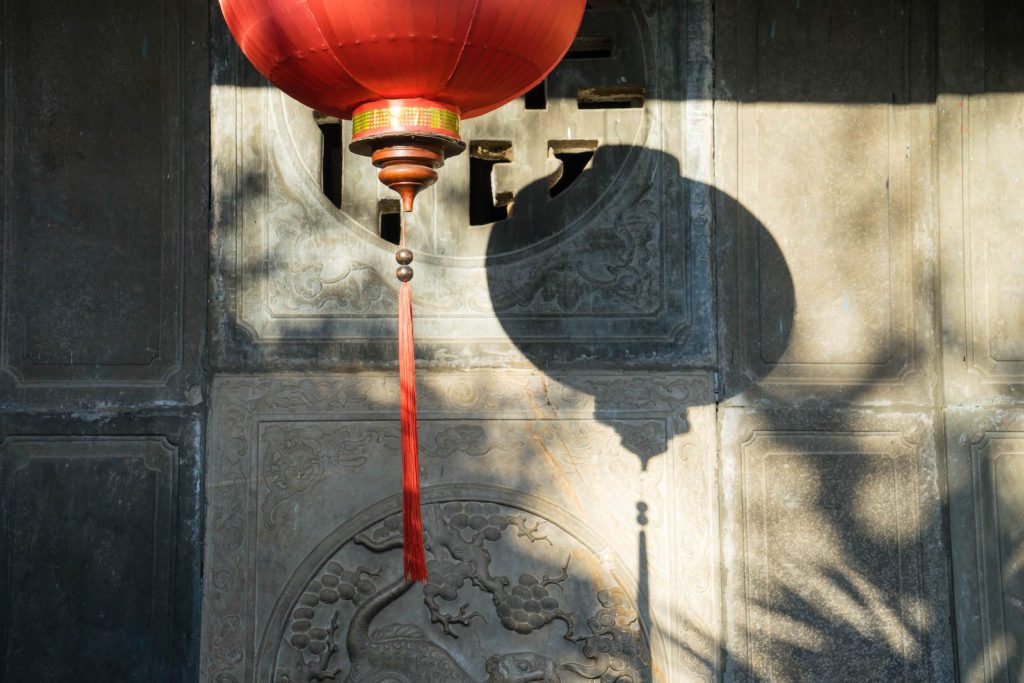 A traditional red Chinese lantern hangs against a grey wall with intricate carvings, casting a detailed shadow in the warm sunlight.