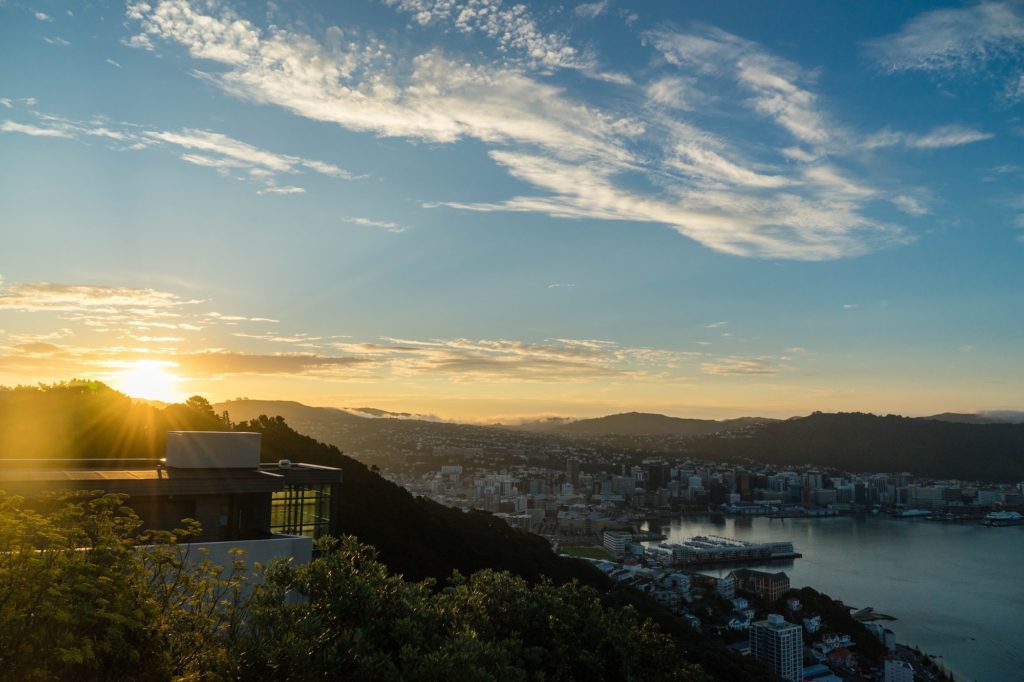 The image depicts a sunrise over a city, showing a waterfront, buildings, and hills, with the sun peeking over the horizon and scattered clouds.