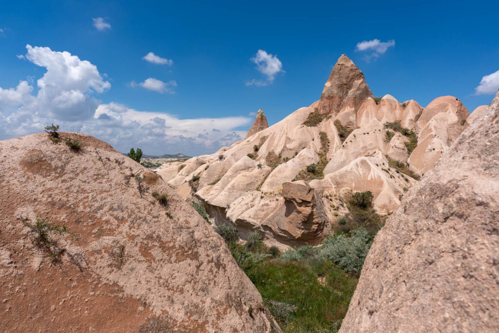 This image features a landscape with unique rock formations, likely from Cappadocia, Turkey, under a blue sky with scattered clouds and sparse vegetation.