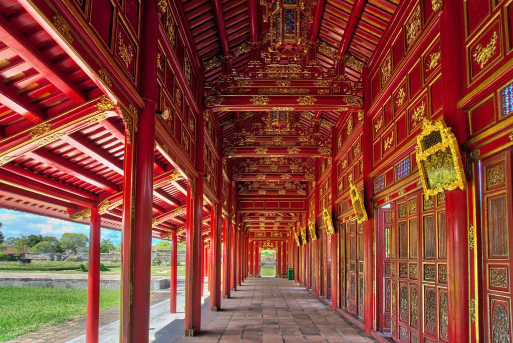 An ornate corridor with red and gold decorations, intricate designs, and wooden columns. Clear skies and greenery are visible outside. The architecture suggests Asian heritage.