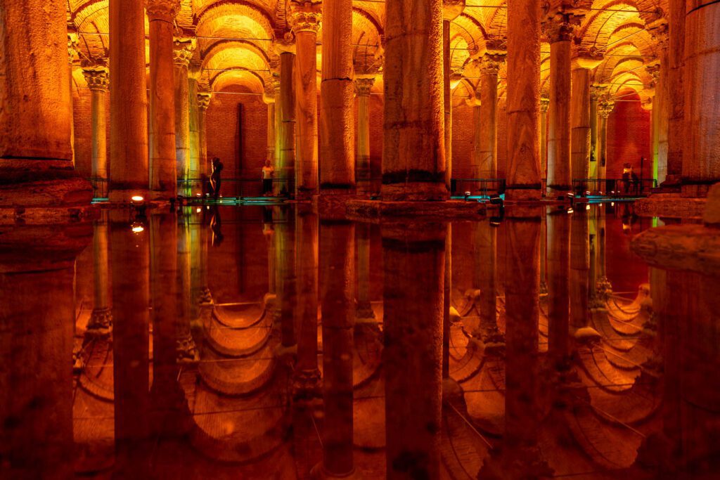 This image shows the interior of an ancient cistern with illuminated columns reflecting on the calm water surface, creating a symmetrical, serene, almost mystical ambiance.