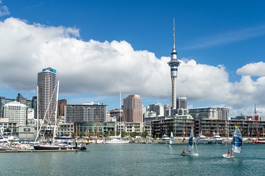 The image shows a scenic waterfront with sailboats, a marina, a tall tower, and modern buildings against a backdrop of blue sky with scattered clouds.