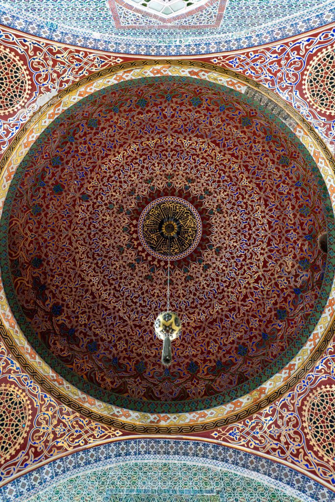 This is an intricately patterned dome ceiling with ornate Islamic geometric and arabesque motifs, featuring vibrant colors and a hanging chandelier.