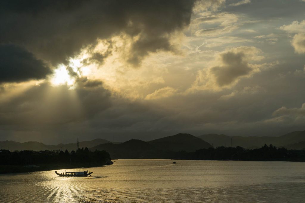 The image features a serene lake with a boat sailing, sunbeams piercing through clouds above mountain silhouettes, and a tranquil, golden-lit water surface.