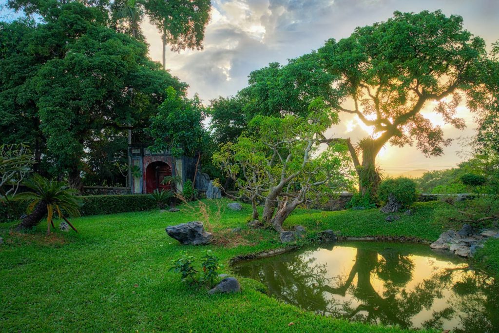 A serene garden at sunset with lush trees, a reflective pond, a stone pathway, and a quaint house with a red door surrounded by greenery.