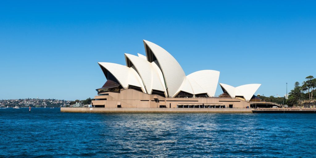 The image shows the Sydney Opera House with its iconic sail-like design on a sunny day, set against the clear blue sky, overlooking the harbour.