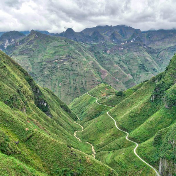 A winding road cuts through lush, terraced green mountains under a cloudy sky. The landscape appears remote and agricultural, with a sense of tranquility.