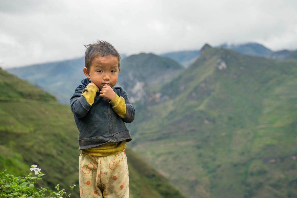 A child stands in front of a mountainous landscape, wearing casual clothes, with a contemplative gaze, and hands near the mouth.