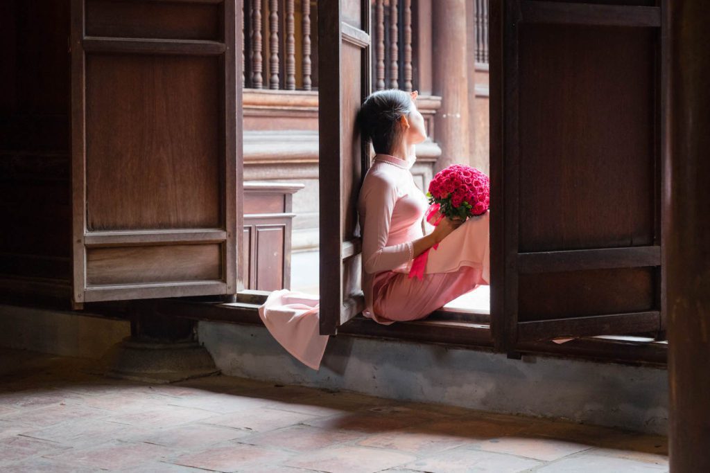 A person in traditional attire sits by an open wooden window, holding a bouquet of red flowers, gazing outside thoughtfully in a serene atmosphere.