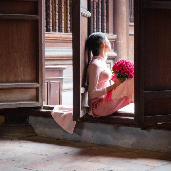 A person in a traditional pink outfit kneels by an open wooden door holding a red bouquet, looking outwards contemplatively in a serene, historical setting.