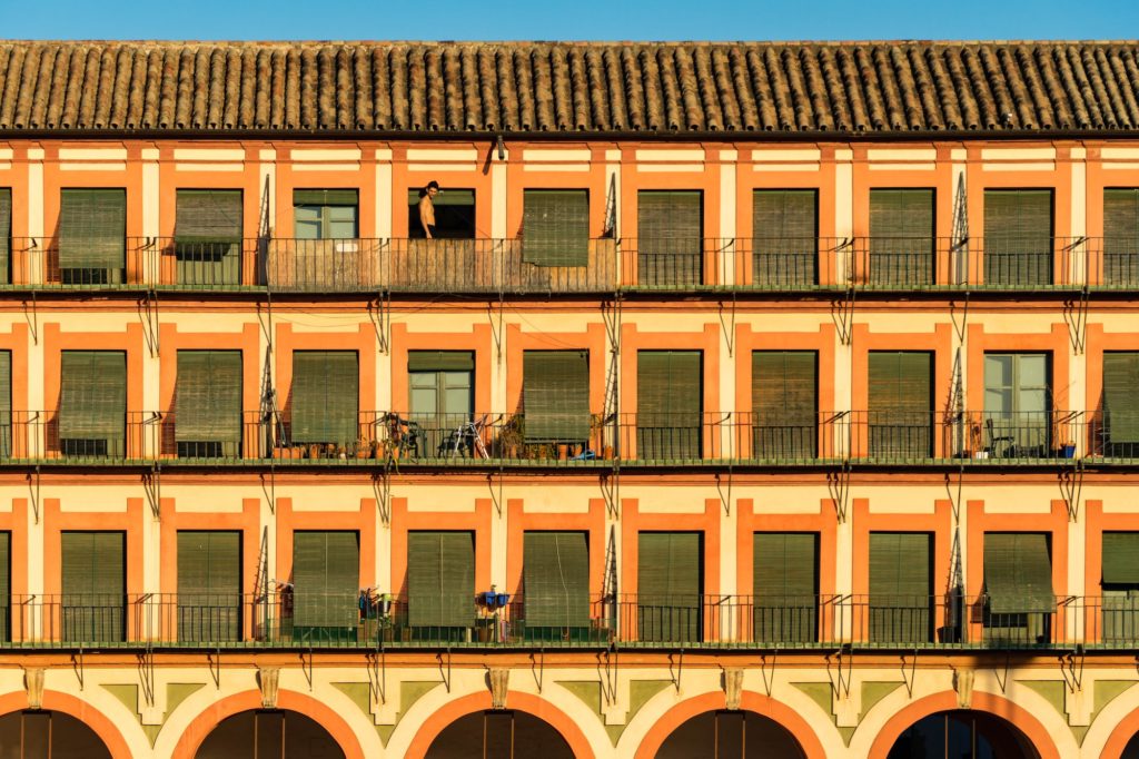 This image shows a symmetrical facade of an orange building with white arches, two levels of balconies, and a terracotta-tiled roof against a clear sky.