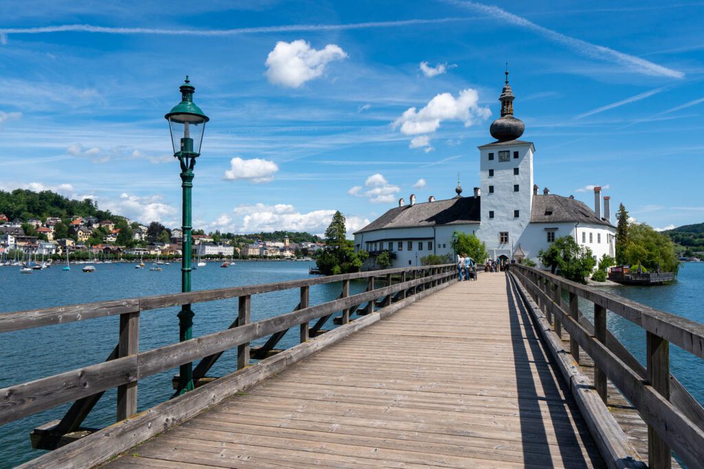 A wooden bridge with a lamppost leads to a white building with a tower against a blue sky. People stroll along, and boats line the water nearby.