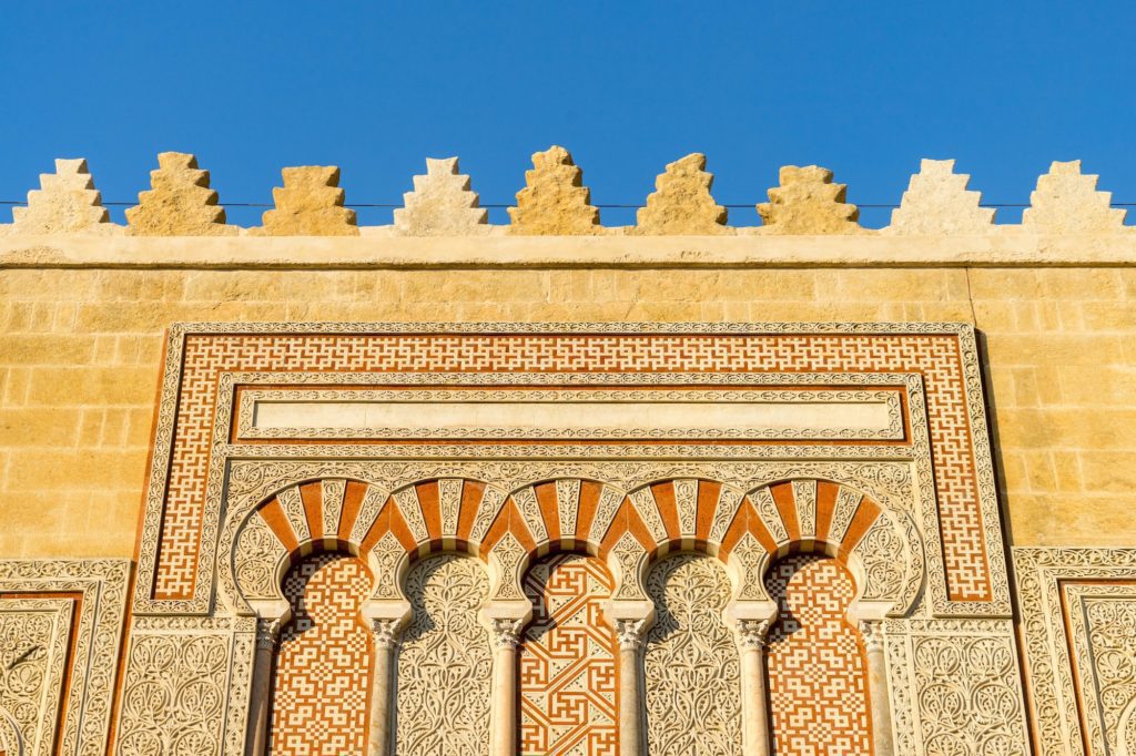 The image displays intricate Islamic architecture with detailed arabesque patterns and muqarnas above arched windows against a clear blue sky.