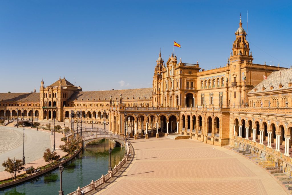 This image shows a grand, ornate building with arches surrounding a plaza featuring a canal and bridge. The Spanish flag flies atop, under a clear sky.