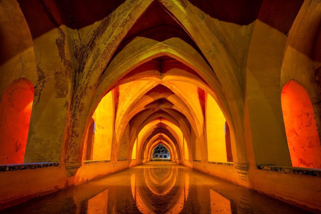 An architectural picture showing a symmetrical view of arched corridors with water reflecting the vivid orange illumination, suggesting a serene, historical, or cultural setting.