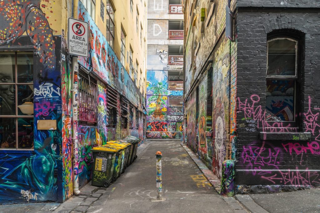 An urban alleyway covered in colorful graffiti. Walls and dumpsters feature vibrant street art, with a no parking sign visible near the entrance.