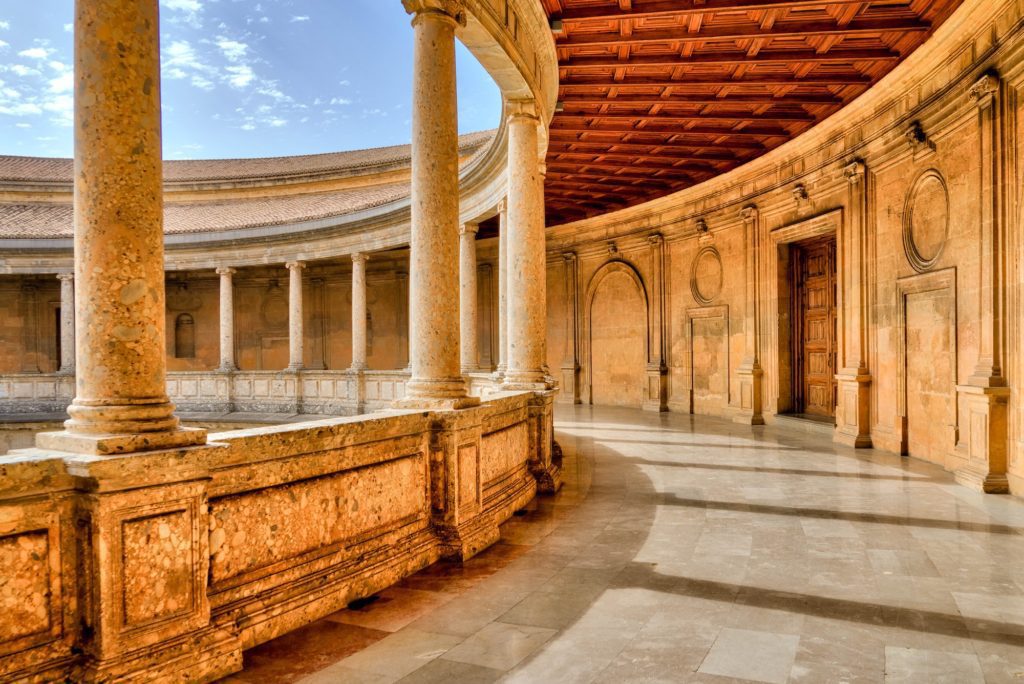 An ancient colonnade with a series of rounded arches, weathered stone balustrades, and a wooden ceiling. Sunlight bathes the elegant architecture and patterned floor.