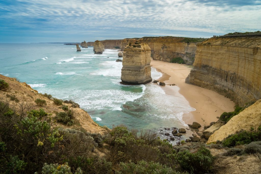 The image shows a breathtaking coastal landscape with rugged cliffs, sandy beach coves, and towering rock stacks against a cloudy sky. Waves crash gently below.