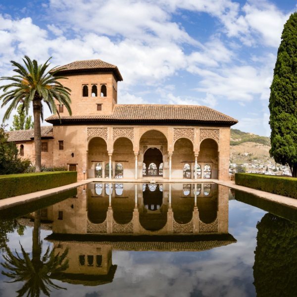 An Andalusian style building with arched porticos, reflected in a tranquil pool, flanked by tall trees under a partly cloudy sky. It's serene and architecturally distinct.