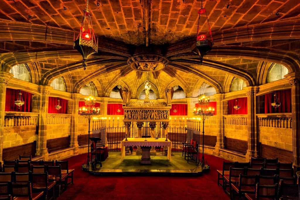 This is an opulent medieval-style hall with arched ceilings, intricate stonework, vibrant red banners, old wooden chairs, and ambient yellow lighting from lanterns.