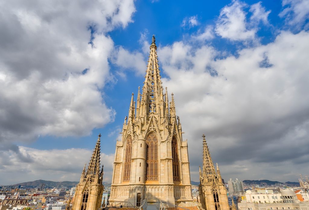 Gothic cathedral spires reach into the sky above a cityscape, with intricate architectural details and statues. Fluffy clouds float in a bright blue sky.