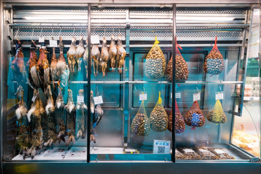A refrigerated display case with various hanging game birds and cured meat cones. The glass doors reflect light, and price tags are visible beneath the items.