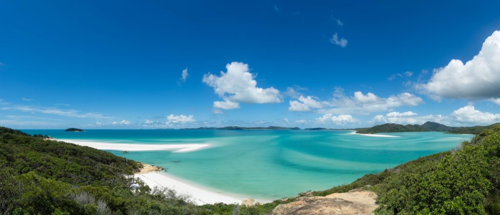 A panoramic view of a serene beach with crystal-clear turquoise water, white sand, lush greenery, scattered clouds above, and distant islands on the horizon.