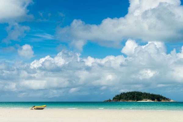 A tranquil beach scene with a solitary yellow boat on white sand, a small island in the distance, under a blue sky dotted with fluffy clouds.