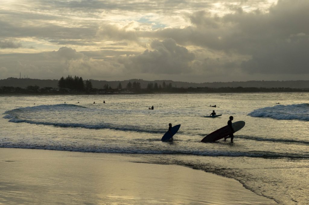 Sunset at a beach with silhouettes of people carrying surfboards and wading into the water, with others surfing waves in the background. Cloudy skies above.