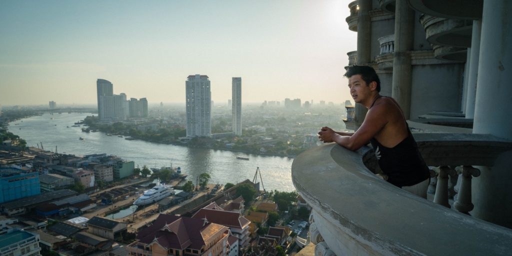 A person sits on a high balcony overlooking a river with high-rise buildings in the distance, during the early hours of sunrise or sunset.