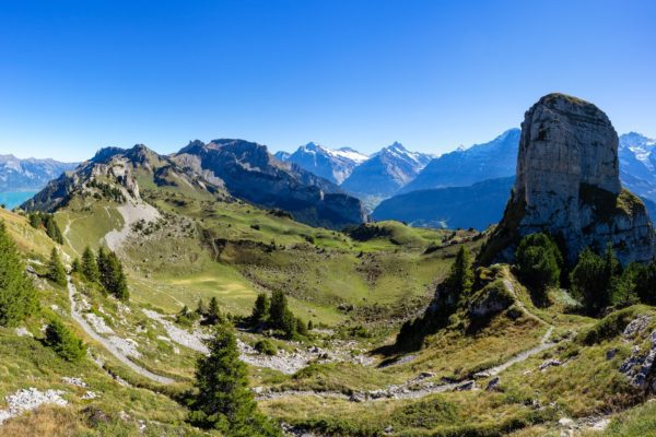 A panoramic view of a lush mountain landscape under a clear blue sky, featuring rugged peaks, green valleys, and a prominent rocky outcrop.