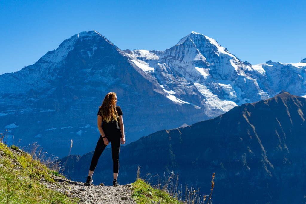 A person stands on a mountain trail overlooking the majestic snow-capped peaks under a clear blue sky, offering a sense of adventure and serenity.