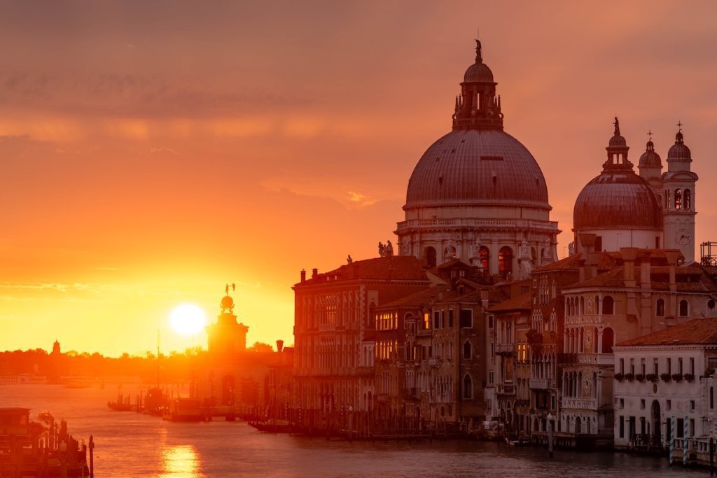 The image captures a stunning sunset in Venice with an orange sky, illuminating the domed Santa Maria della Salute church and the Grand Canal.