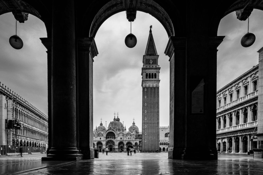 This black and white image captures a view of St. Mark's Square in Venice from under an archway, featuring the campanile and basilica.