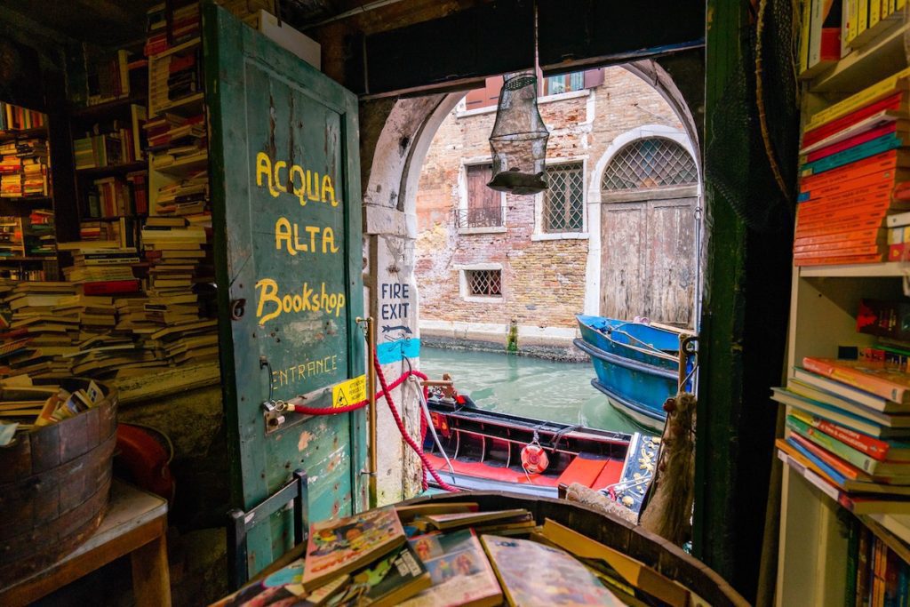 A cozy bookshop, Acqua Alta, with an open door leading to a canal where a red boat is moored. Shelves with books line the walls inside.
