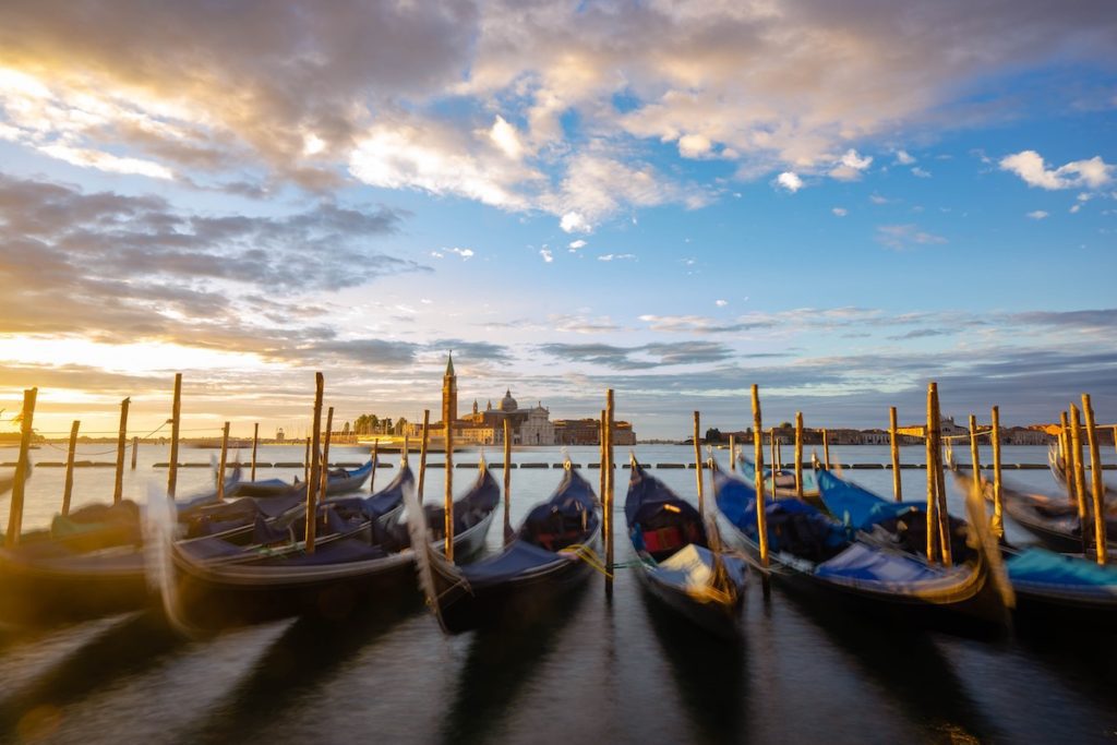 A beautiful sunset with golden and blue hues over a serene lagoon where multiple gondolas are moored to wooden poles, with an historic building in the background.