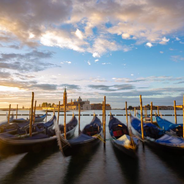 Gondolas moored on gently rippling water at sunset with a historic building in the backdrop under a sky streaked with clouds in Venice, Italy.