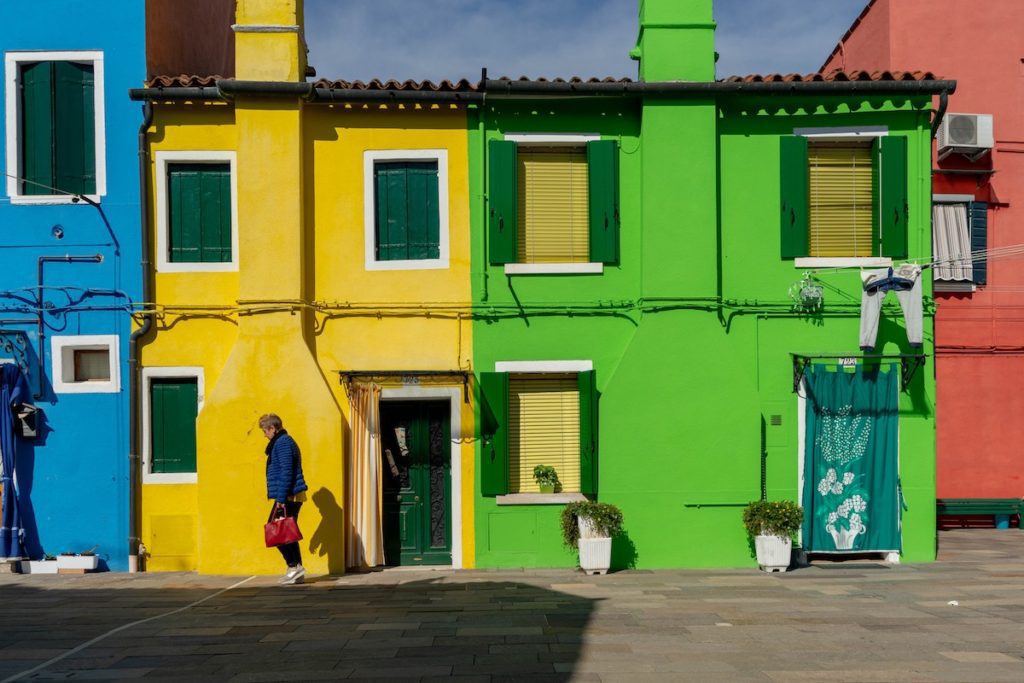 A person walks by brightly colored houses with closed shutters and laundry hanging outside under a clear blue sky.