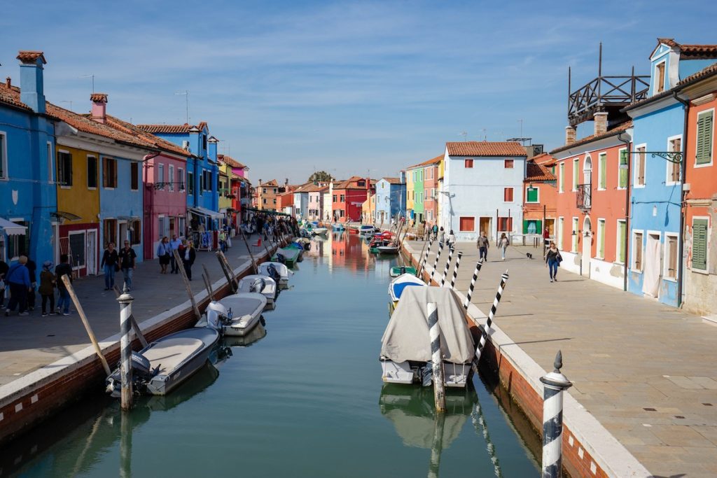 A canal lined with colorful houses under a clear sky. Boats are moored alongside, and people stroll beside the water. It resembles Burano, Venice.