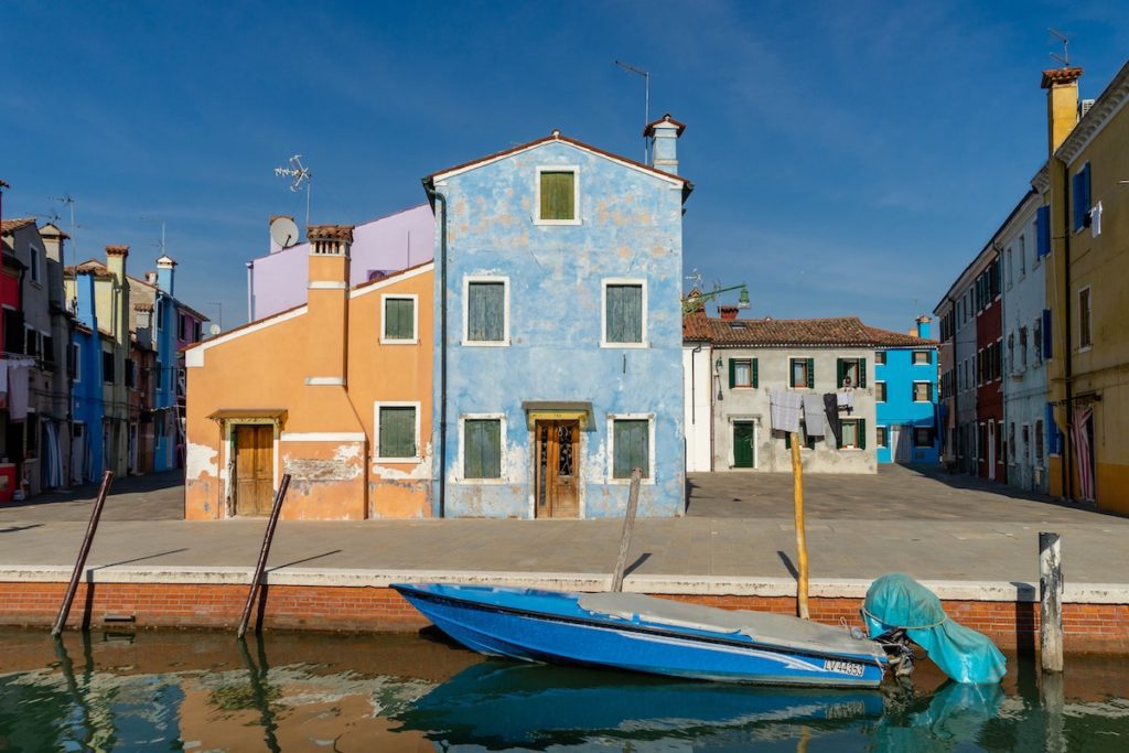 A picturesque canal in Burano, Venice, with colorful plastered buildings, a clear blue sky, and a small boat moored to a stone dock.
