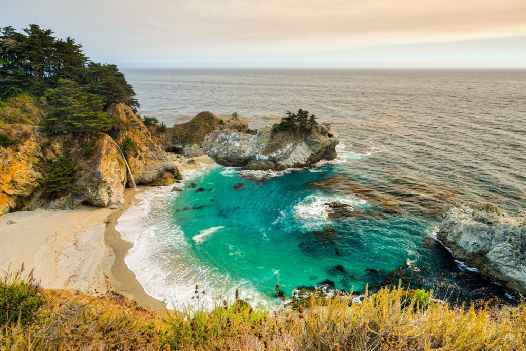 A serene coastal scene with turquoise waters, sandy beach, rugged cliffs, and greenery under a hazy sky, suggesting a peaceful and natural vista.