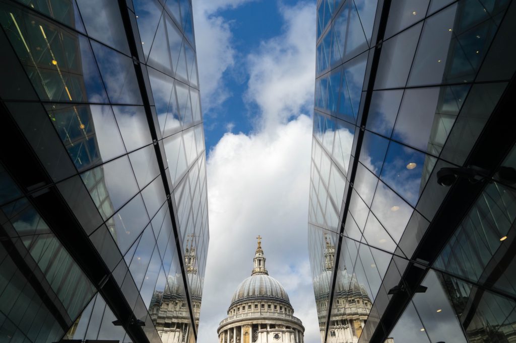 The image shows a symmetrical view of a cathedral dome framed by reflective glass buildings, under a bright sky with scattered clouds.