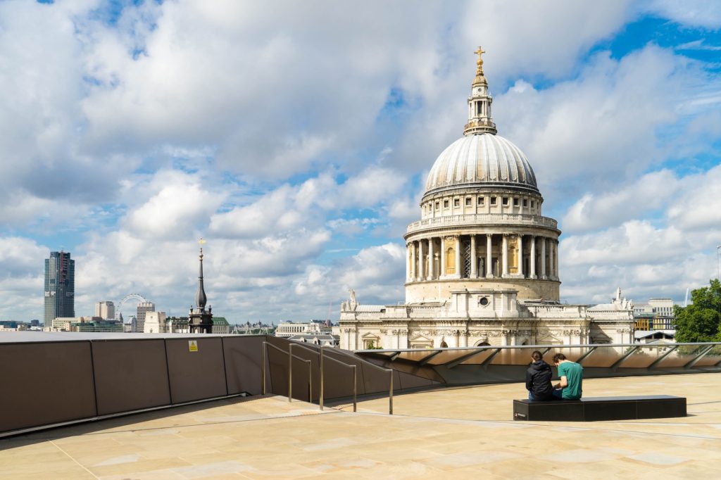 The image shows St. Paul's Cathedral in London. A person sits on a bench, gazing at the iconic dome under a partly cloudy sky.
