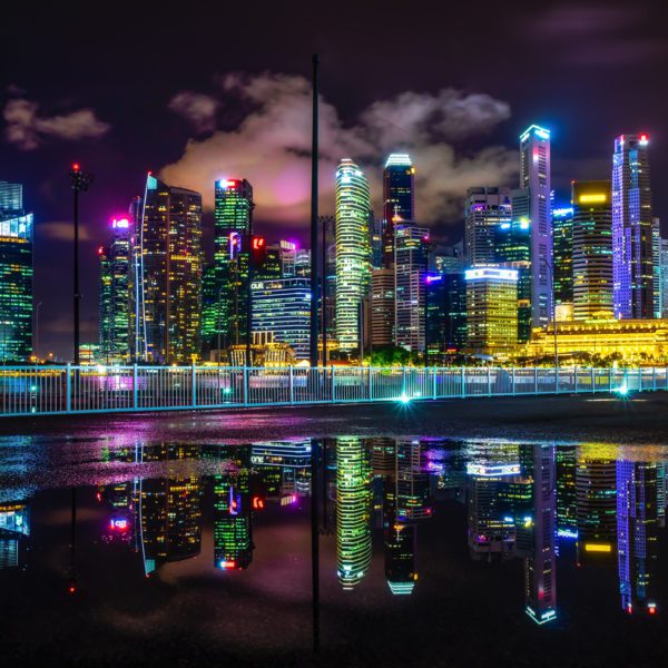 A vibrant cityscape at night with illuminated skyscrapers reflected on a smooth water surface. Colorful lights contrast with the dark sky above.